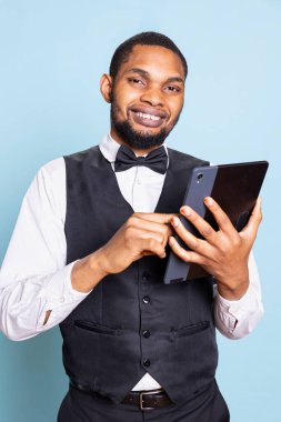 Smiling hotel concierge reviewing all online room reservations on tablet, checking the resort website against blue background. Doorkeeper employee managing bookings in studio. clipart