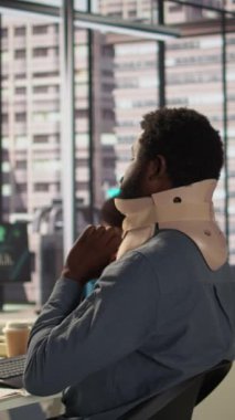 Servikal tasmalı dikey Video Hurt çalışanı yeni bir iş projesi üzerinde çalışıyor, boynu kırılmış bir şekilde masada oturmaya çalışıyor. Afrikalı Amerikan şirketinin ortağı görevleri çözmeye çalışıyor