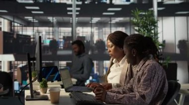 Afrikalı Amerikalı iş kadınları aylık gelir dengesi hesaplamalarında işbirliği yapıyor, girişim geliştirme planı hazırlıyorlar. İdari personel ekibi bilgisayardaki analizleri değerlendiriyor. Kamera B.