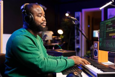 Profesyonel yazılımda akustik kaydetmek ve ses efektleri eklemek için elektronik klavye çalan Afrikalı Amerikalı bir besteci. Ses mühendisi stereo konsol ve araçlarla ses parçalarını düzenliyor.