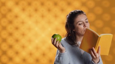 Kitap okuyan ve taze elmanın tadını çıkaran, sağlıklı yaşayan, stüdyo arka planında izole edilmiş bir kadın. Yeşil meyve yiyen, biyo-atıştırmalıkların tadını çıkaran bir kitap kurdu, kamera A