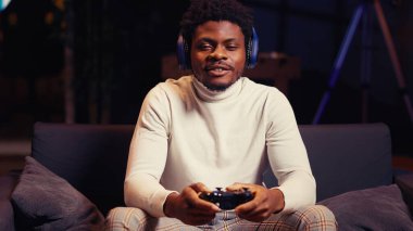 Afrikalı Amerikalı bir adam arkadaşlarıyla çevrimiçi oyun oynuyor, kulaklıkla konuşuyor. Oyun kurucu, oyun alanı denetleyici kullanarak oyun konsolunda takım arkadaşlarıyla video oyunları oynuyor