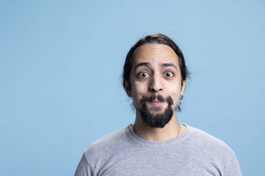 Uzun kıvırcık saçlı, stüdyoda kendine güvenen bir erkek model mavi arka planda üstünkörü giyiniyor. Kendine güveni tam barışçıl genç bir adam..