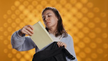 Sırt çantasını karıştıran, kitap çıkaran, arka planda izole edilmiş bir kadının portresi. Okul çantasından eğitim amaçlı kullanılan ders kitabını kaldıran öğrenci, kamera B