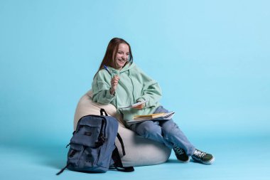 Armut koltuğa oturmuş gülen neşeli kızın portresi ve defter, stüdyo arka planı. Kollarında not defteriyle aşırı neşeli liseli kız ödevlerini hazırlarken eğleniyor.