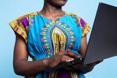 Klasik Afro-Amerikan kıyafeti giymiş genç bir kadın stüdyoda dizüstü bilgisayar tutuyor, web sayfalarını ve sosyal medya platformlarını keşfetmek için internette sörf yapıyor. Gülümseyen, modern kadın kablosuz internet teknolojisinden hoşlanıyor..