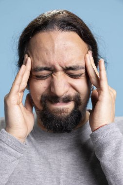 Stresli yetişkin, mavi arka planda baş ağrısıyla boğuşuyor, şakaklarını ovuyor ve yorgunluk ya da anksiyeteden dolayı ağrısını azaltmaya çalışıyor. Genç adamın migreni ve yorgunluğu var..