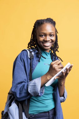 Okul sınavı için gülümseyen ve yazı yazan mutlu bir kızın portresi not defterinde, stüdyo arka planında. Neşeli sırıtan öğrenci, üniversite dersleri için gerekli araştırmaları tamamlıyor.