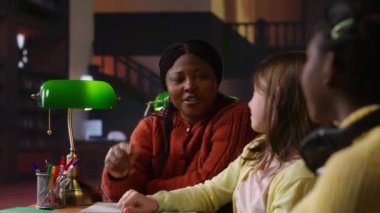 İlkokul öğrencileri bir profesörle matematik çalışıp akademik becerilerini kütüphane masasında geliştiriyorlar. Siyahi kadın müfredat materyallerini açıklıyor. Kamera B.