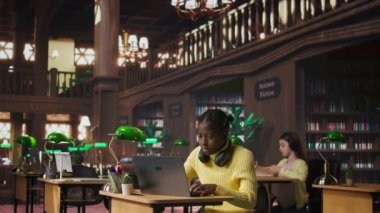 Afrikalı Amerikalı liseli bir kız kütüphanede dizüstü bilgisayara ders notları yazıyor, bir çalışma masasında online bilgi içeren bir proje üzerinde çalışıyor. Öğrenci akademik enstitüde ders için hazırlanıyor. Kamera B.