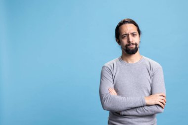 Ciddi bir insan kamerada hayal kırıklığı yaratan yüz ifadeleri yapıyor, kollarını mavi arka planda kavuşturup düşünüyor ve poz veriyor. Stüdyoda özgün bir erkek model..