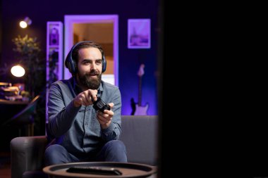 Joyous man having fun while playing videogames on gaming console with teammates, talking through headphones. Lively gamer chatting with friends through headset while defeating players online clipart