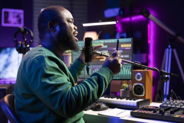Afrika kökenli Amerikalı besteci, ses kalitesini arttırmak için özel efektler ekleyerek ve miksaj konsoluyla düzenleme yaparak şarkısını kaydediyor. Erkek sanatçı söz yazarı mikrofonla şarkı söylüyor..