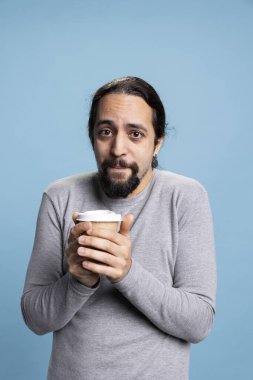 Rahat bir insan mavi arka planda bir fincan kahve içerek stüdyoda kafein servisi yapar. Genç yetişkin soğuk ve kendinden emin hissederken taze, siyah bir içecek içiyor..