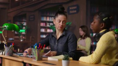 Kütüphane masasında ilkokul kızına rehberlik eden beyaz bir öğretmen, akademik gelişimlerine yardımcı olmak için çalışma kitapları ve notlarla özel dersler veriyor. Öğretmen akademisyenle konuşuyor. Kamera B.