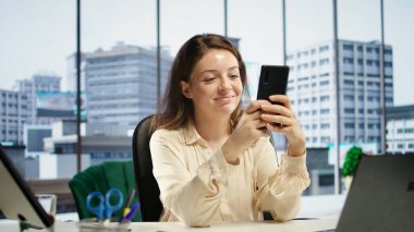 Pleased empowered businesswoman texting messages in her own director office, having fun on social media. Leader discussing with partners about new project ideas for career growth. Camera B. clipart