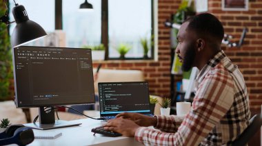 Genç bir adam, şık bir ev ofisinde dizüstü bilgisayarında yazılım kodları yazarken görülmüş. Çift monitörlü çalışma alanı yazılım mühendisliği, web geliştirme ve kodlama verimliliğine yardımcı olur. Kamera B.