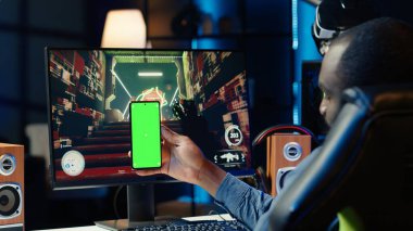 Man texting friends on green screen phone while playing first person shooter videogame at home. Gamer looking at mockup smartphone display, enjoying online discussion with mates clipart