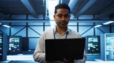 System administrator in data center using notebook to design, implement and manage gear, ensuring optimal performance and reliability. Indian man in server room coding on laptop, camera B clipart