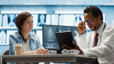 Afro-amerikan sıhhiyeci kadın bir hastaya teşhis raporlarını sunuyor, tıbbi muayeneden sonra laboratuvar sonuçlarını gösteriyor. Doktor profesyonel tavsiye veriyor. Kamera A.