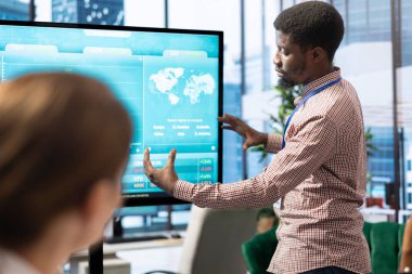 Genel müdür interaktif panoda bilgi grafikleri ve satış numaralarıyla bir sunum yapıyor, personele organizasyonel ilerlemeyi gösteriyor. Çalışanlar analiz planlarını gözden geçiriyorlar.