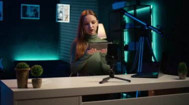 Etkileyici, taşınabilir hoparlör performansını değerlendiriyor, kullanılan parçalardan bahsediyor, telefonla çekim yapıyor. Tripodun üzerinde akıllı telefon kullanan kız müzik cihazı incelemesi, kullanılmış malzemelerden memnun, kamera B