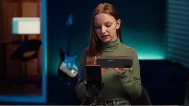 Etkileyici, taşınabilir hoparlör performansını değerlendiriyor, kullanılan parçalardan bahsediyor, telefonla çekim yapıyor. Tripodun üzerinde akıllı telefon kullanan kız müzik cihazı incelemesi, kullanılmış malzemelerden memnun, kamera A