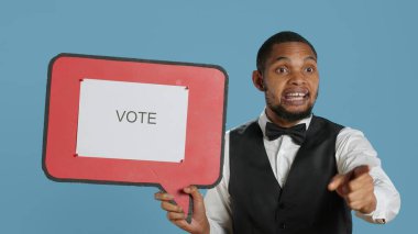 Political bellboy hotelier showing a speech bubble with the message to vote, advertising politics for a marketing commercial. Hotel porter staff in tuxedo holds a cardboard icon. Camera A. clipart