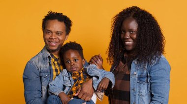 Stüdyoda sarı arka plana gülümseyen Afro-Amerikan ailesi sevgi ve içten şükranla poz veriyor. İyimser anne ve baba bebeğini kucağında tutuyor, birlikte vakit geçiriyorlar. Kamera A.