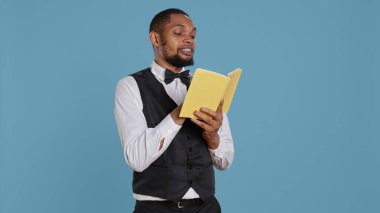 Genç kapıcı, stüdyo molasında kitap okuyor. Smokinli otel görevlisi, mavi arka planda. Belboy tatil köyü personeli edebiyat ve konferansı sever. Kamera B.