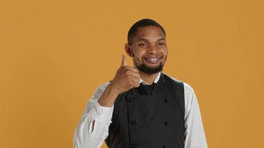 Waiter smiling and showing a thumbs up symbol against yellow background, presenting his satisfaction and approval with a positive like sign. Cheerful staff gives okay gesture. Camera A. clipart