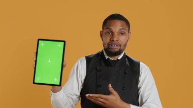 Restaurant waiter presenting a green screen layout on tablet against yellow background, wearing professional service oriented attire on duty. Butler serving clients, professionalism. Camera A. clipart