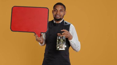 Catering employee showing a red speech bubble icon in studio, carrying a cruet set with condiments. Waiter with professionalism presenting an empty cardboard sign as an advertisement. Camera B. clipart