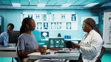 Siyah sıhhiyeci tansiyon monitörüyle kan basıncı ölçer, kontrol için modern gelişmiş aletler kullanan kadın hastalarla konsültasyon yapar. Kamera B.