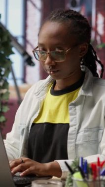 Ofisteki neşeli kadının dikey video portresi dizüstü bilgisayarı açıp görevler üzerinde çalışmaya başlıyor. Neşeli Afro-Amerikan vatandaşı elektronik not defteri kullanarak online iş yapıyor, kamera B