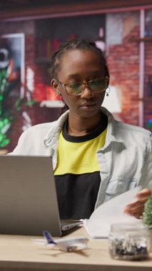 Ev ofisinin masasında, dizüstü bilgisayarla dosya inceleyen ve sözleşmeler hazırlayan dikey video uzaktan kumanda işçisi. Uzaktan çalışan, defter, kamera B önündeki evrakları kontrol ediyor.