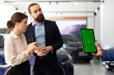 Araba galerisi çalışanı elinde bir model cihaz tutuyor, müşterilerine istenen aracı belirlemek için sorular soruyor. Çalışan müşterilerin memnuniyetini sağlar, sorulara cevap verir, yeşil ekran tabletinde arabaları gösterir