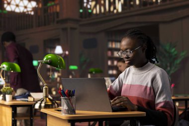 Üniversite kütüphanesindeki genel veritabanından veri toplayarak bir edebiyat ödevine odaklanan genç zenci öğrenci, projesi için daha fazla akademik bilgi toplamak için dijital araçlar kullanıyor..