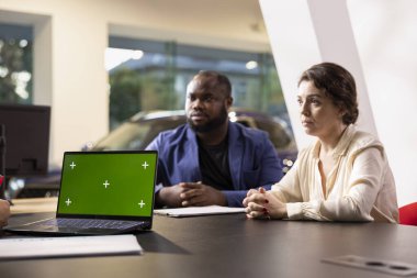 Satıcı müşterilere yeşil ekran dizüstü bilgisayarda araba özelliklerini gösteriyor, anlaşmaya varmayı umuyor. Showroom temsilcisi müşterileri satın almaya ikna etmeye çalışıyor. Aracın eklentilerini maket defterinde sunarak.