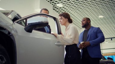 Araba satış temsilcisi müşterileri araç kapısına ve test kalitesine dokunmaya davet ederek bireylerin yatırım kararı almalarına yardımcı oluyor. Showroom temsilcisi müşterilerden otomobil özelliklerini kontrol etmelerini istiyor, kamera B