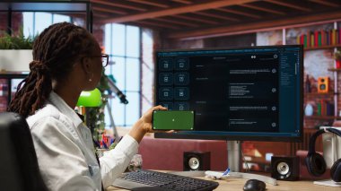 AI LLM sohbet robotu yazılımını takip etmek için izole ekran cep telefonu kullanan bir kadın. Afrikalı Amerikalı apartman dairesinde akıllı telefon ve bilgisayar ve kamera A da GPT programı kullanıyor.