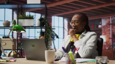 Ofisteki telgraf işçisi dizüstü bilgisayarla çalışırken esniyor ve yorgun hissediyor. Yeşim Afro-Amerikan uzaktan kumandalı çalışan rahatlayan kaslar, not defterindeki görevleri çözen, kamera A