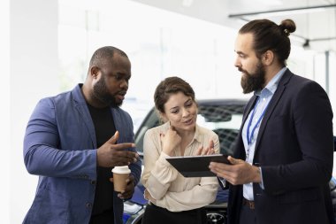 Elinde tablet bulunan satış temsilcisi müşterilere arabalar, finansman ve alım süreci hakkında sorular soruyor. Showroom ajanı, aygıt kullanan müşterilerle garantiler ve hizmetler hakkında konuşur