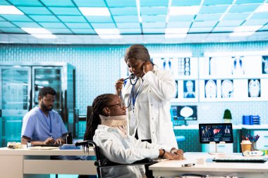 Siyahi hekim steteskop kullanarak hastasına danışmanlık yapıyor. Akciğerleri ve kalp atışlarını dinliyor. Rutin solunum kontrolünde servikal köpüklü tekerlekli sandalye kullanıcısı..