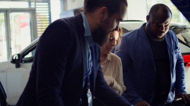 Araba galerisi satıcısı müşterilerle ihtiyaçlarını tartışmak için buluşuyor, araç kaputu altında bileşenleri gösteriyor. Otomobil özelliklerini müşterilere sunan gösteri odası temsilcisi, kamera B