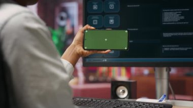 Ana ofisteki bilgisayar ekranında AI LLM sohbet robotunun nasıl kullanılacağını öğrenmek için yeşil ekran akıllı telefonu kullanan bir kadın. Kişi, yapay zeka sohbet robotları ve kamera B hakkındaki video dersini model telefondan izler.
