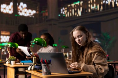 Genç akademisyen, kampüs kütüphanesindeki sınav hazırlıklarına dikkatle odaklanıyor, laptopuna barışçıl bir çalışma ortamındaki referans kitaplarından notlar alıyor. Kız genel veritabanı materyallerini kullanıyor.