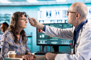 Genel pratisyen hekim termometrenin ateş ölçümlerini alıyor, kabinedeki yaşlı kadın hastaya danışıyor. Yaşlı doktor hastalık önleme sıcaklığını kontrol ediyor..