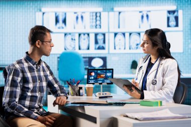 Uzman ve hasta modern bir muayenehanede teşhis sonuçlarını tartışıyor, röntgen görüntüleme sonuçları ve MRI taramaları sunmak için gelişmiş sağlık teknolojisi kuruluyor. Bakım yönetimi.