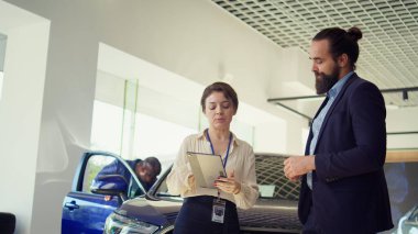 Satıcı kadın, araba seçimini erkeklere göstermek için tablet kullanıyor. Satın alması için onu ikna etmeye çalışıyor. Showroom temsilcisi müşteriye anlaşma öneriyor, aygıtta ayrıntıları gösteriyor, satış taktikleri kullanıyor, kamera B